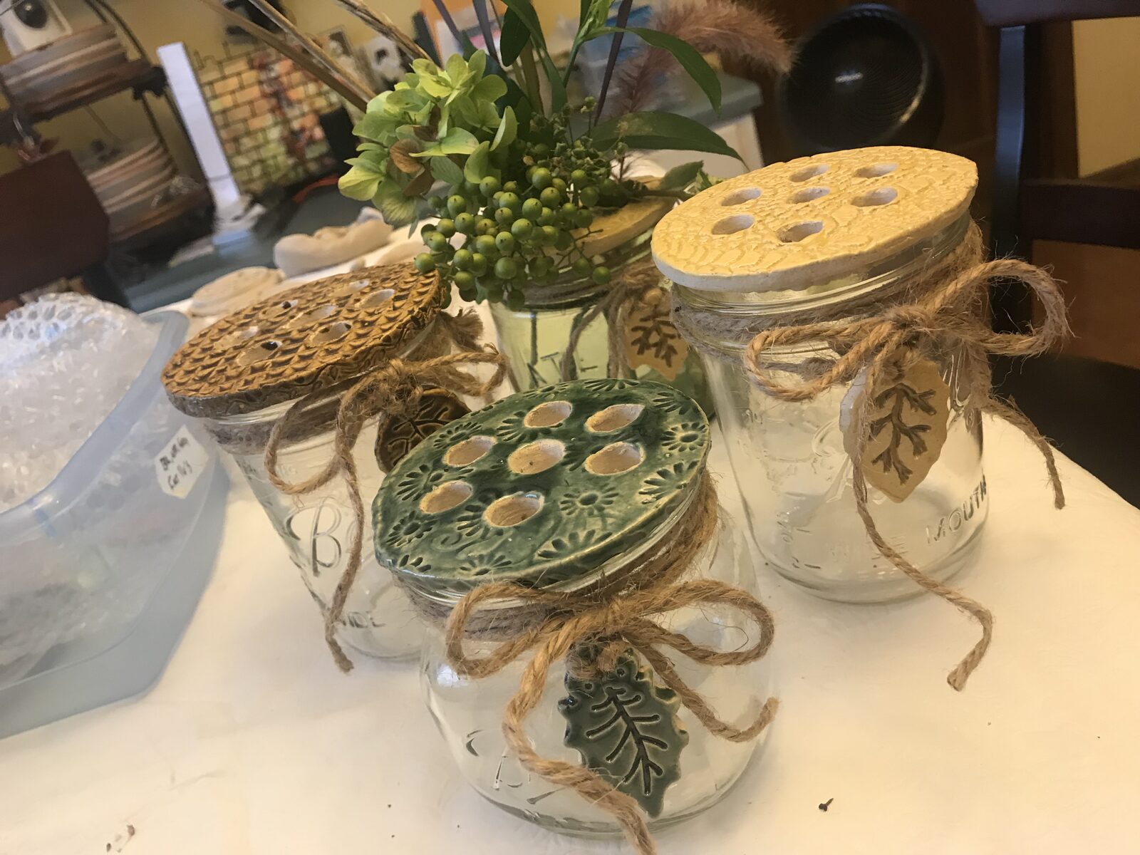 Mason jars topped with ceramic flower frog lids in yellow, brown, and green glazes, tied with twine and leaf charms