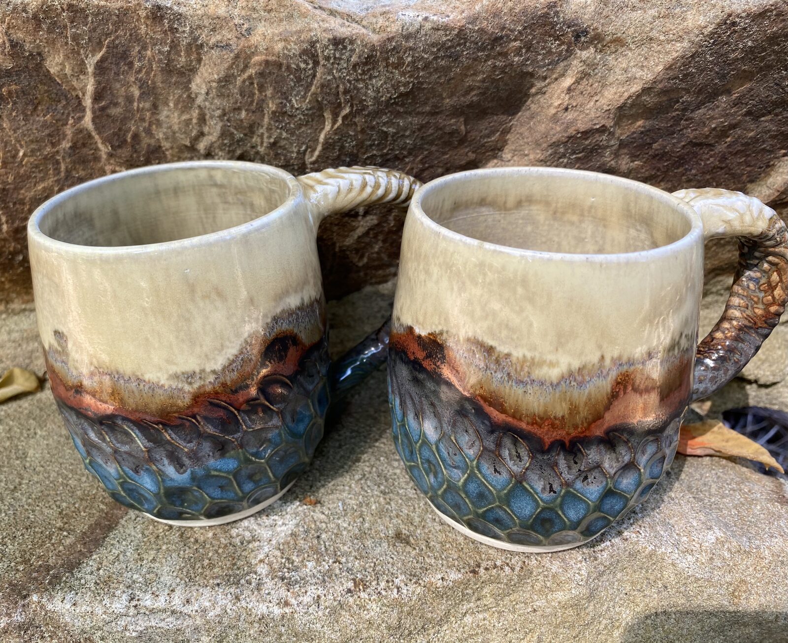 Pair of mugs with cream tops, copper drip bands, and textured honeycomb-blue bottoms with twisted vine handles
