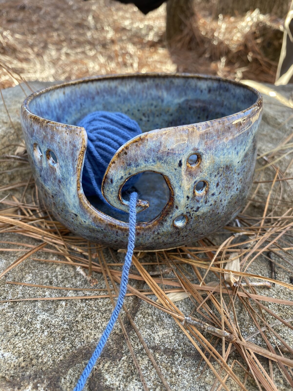 Yarn bowl with curved cutout for guiding yarn, holding a ball of blue yarn, in speckled lavender-blue glaze